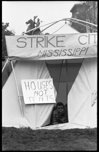 Man lying his tent, looking out the front flap: signs reading 'Strike City Mississippi' and 'Houses not tents,' statue of Lafayette in the background