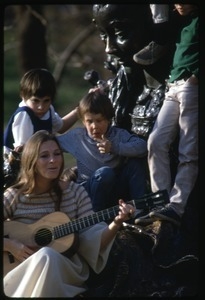 Judy Collins seated at the base of a statue, playing guitar for children