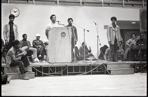 Huey P. Newton speaking at Boston College: Newton at podium, surrounded by Party members, including David Hilliard (seated under clock)