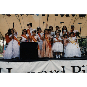 Women and girls in dresses and sashes pose and wave on stage at the Festival Puertorriqueño