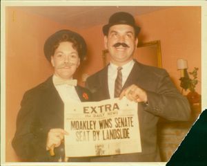 John Joseph Moakley and Evelyn Moakley, wearing suits and fake mustaches, holding newspaper with headline "John Joseph Moakley Wins Senate Seat By Landslide," 1960s