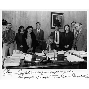 Mayor Menino at his desk, signing papers, surrounded by a group of people that includes Clara Garcia.