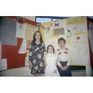 Three children posing in front of a display of children's drawings and stories.