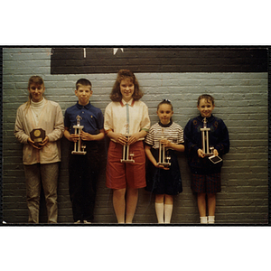 A boy and four girls in a line pose against a wall, holding their trophies
