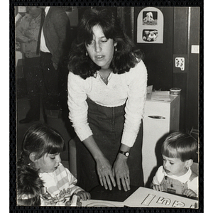 An Unidentified teacher observes a group of children at a table working on an art project