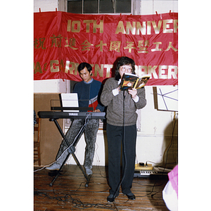 Facing front, a woman sings from a book while a man accompanies her on the keyboard at the Association's tenth anniversary celebration