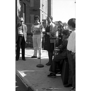 Woman speaks into microphone holding a rolled sheet of paper at a Latino street festival, with onlookers listening and musicians waiting to play music