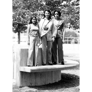 Three young Hispanic women standing on top of a bench, outside, at a La Alianza retreat, at an unidentified campus, [Aug.?] 1978.