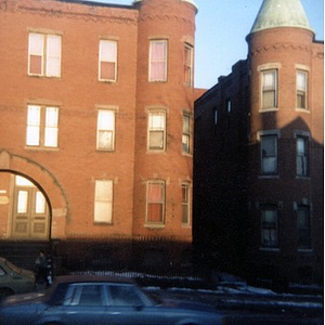 Street scene showing the exterior of two brick apartment buildings and a car driving by in Roxbury.