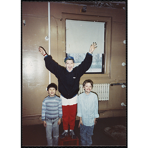A boy standing on a red plastic crate and raising his arms in the air while two other boys stand on each side of him at the Boys & Girls Club