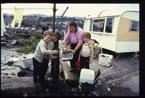 Travellers in Newry doing their washing at a stand up tap, two traveller children looking out through a caravan window, traveller mothers with their babies and group of 7 traveller children, one holding rooster (cockerel) in front of INLA graffiti on wall