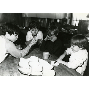 A boy spreads butter on a slice of bread at a table as three other boys look on in a kitchen