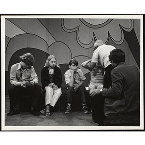 Four teenage girls and boys sit together on the set of a television show [?] while facing a man in the foreground