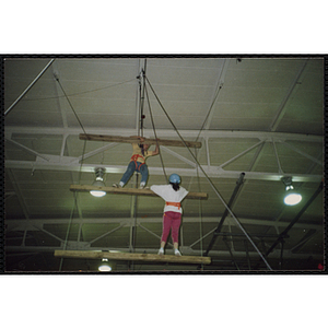 A girl and an indoor climbing supervisor stand on wood beams in a gymnasium