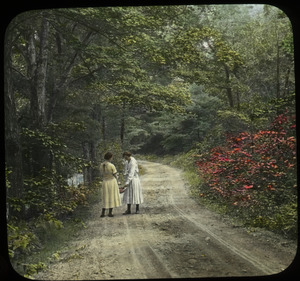 Two women on dirt road bordered by trees and shrubs