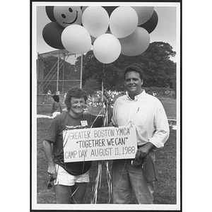 Two staff members pose with "Together we can" sign in front of balloons at Camp Day event