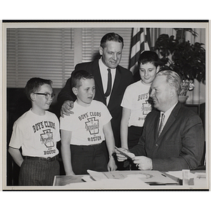 A man sitting at his desk and smiling at a group of three boys wearing Boys' Clubs of Boston t-shirts while William J. Lynch stands behind them