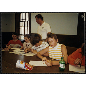 A Group of children writing at a table, while an unidentified man is standing behind them