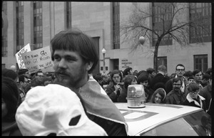 Man in a cape holding a paper mache Nixon mask, standing by a police car during the Counter-inaugural demonstrations, 1969, and march against the War in Vietnam