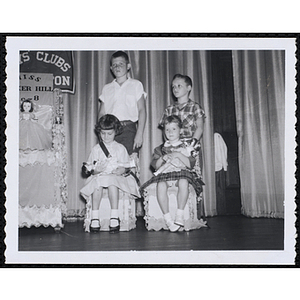 Two winners of the Little Sister Contest sitting in decorative chairs and holding their dolls while their brothers pose behind them