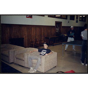 A teenage boy sitting on a couch in the recreation room while others play table tennis at the Charlestown Boys and Girls Club