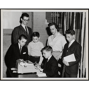 A boy uses a typewriter as three other boys, a woman, and a man look on