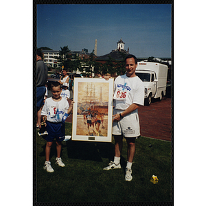 A boy holding a trophy and a man pose with a framed print during the Battle of Bunker Hill Road Race