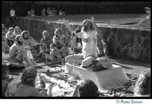 Ram Dass seated on a small platform, arms raised, during his appearance at Andrews Amphitheater, University of Hawaii