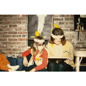 Woman and girl in feathered headdresses and vests sitting together