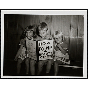 A boy sits with his two sisters, holding a book titled "HOW TO WIN A BEAUTY CONTEST" at a Boys' Club Little Sister Contest