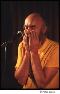 Ram Dass with his hands on his beard, on stage with Amazing Grace