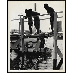 A Youth leaning down over a dock and reaching into the water with a net, while a group of children looks on