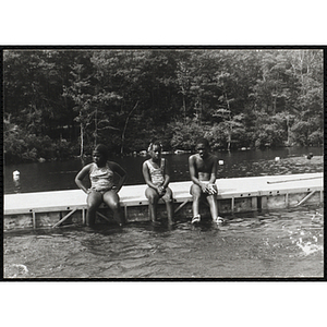 Two girls and a boy sit on a dock with their legs in the water
