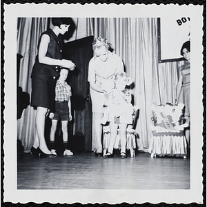 A woman presents a sash to a girl while her brother looks on at a Boys' Club Little Sister Contest