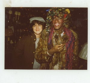 A Photograph of Marsha P. Johnson Wearing a Christmas Lights Headpiece and a Fur Coat, Posing with Another Person at Uplift Lighting
