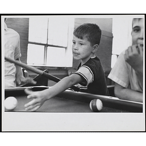 A boy reaches for the cue ball at a pool table