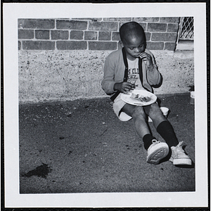 A boy sits against a wall and eats at a picnic