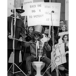 A man sits on a toilet with several people around him at a rally for Mayor of Huntington Avenue