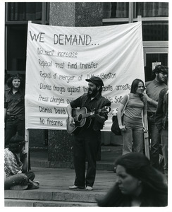 Board of Trustees fee increase demonstration: man playing guitar with protestors on the steps of the Student Union, Charles Bagli holding sign (l)