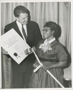 A man holds the 1960 Thumbs Up Award presented to the Ideal Precision Meter Company with a woman holding a forearm crutch