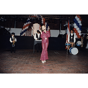 A girl sings into a microphone while three children play instruments in the background