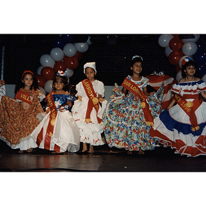 Five girls hold their dresses and wear sashes on stage at the Festival Puertorriqueño