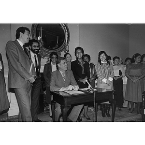 Raymond Flynn sits at a desk with several people behind him