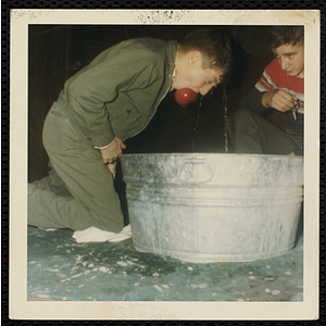 A boy retrieves a bobbed apple from a large basin as another boy looks on