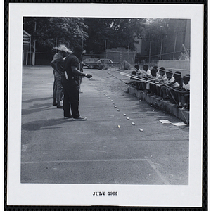 A row of seated boys play a fishing game on a playground as men look on during Tom Sawyer Day