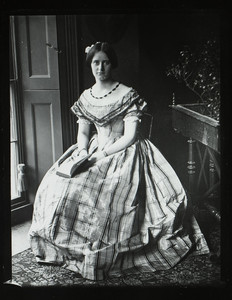 Unidentified young woman, full-length portrait, seated on a chair, facing forward, holding an open book, location unknown, undated