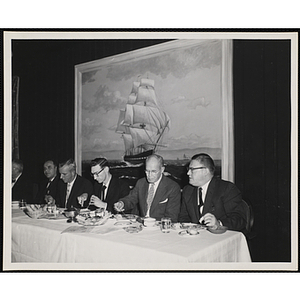 Several men, seated at a long table, eat and converse at the Kiwanis Club's Bunker Hill Postage Stamp Luncheon