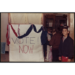 A boy posing next to "VOTE! NOW" sign while others stand in line at the South Boston Boys and Girls Club
