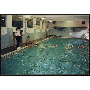 Children stand by and swim in a natorium pool as a woman looks on