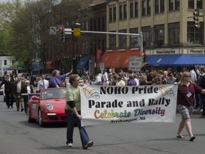 Women marching with 'Noho Pride Parade and Rally' banner, with Mayor Clare Higgins following in a car: Pride Parade; Main Street, Northampton, Mass.
